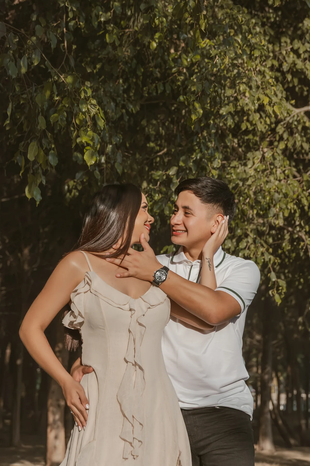 Primer plano de la pareja mirándose y sonriendo bajo los árboles